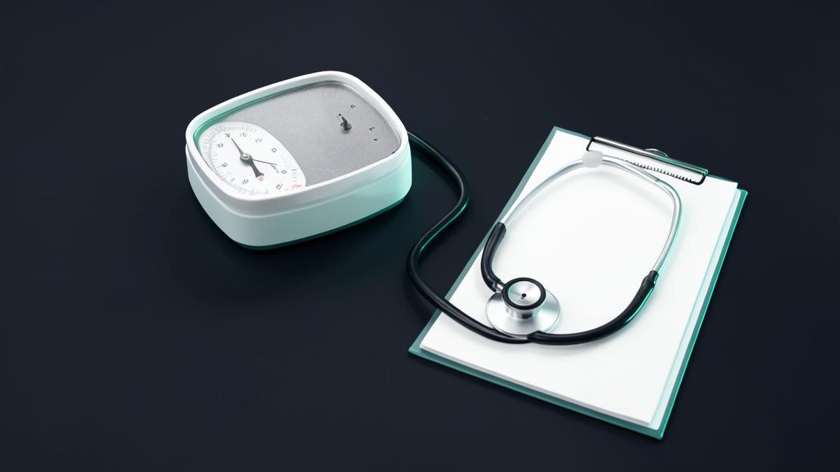 A clinical scale, stethoscope and clipboard on a dark charcoal background with teal accent lighting